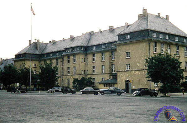Farbfoto der Ray Barracks in Friedberg mit Blick auf das Kasernengebäude, eine aufgezogene US-Flagge und parkende Fahrzeuge aus den 1950er-Jahren